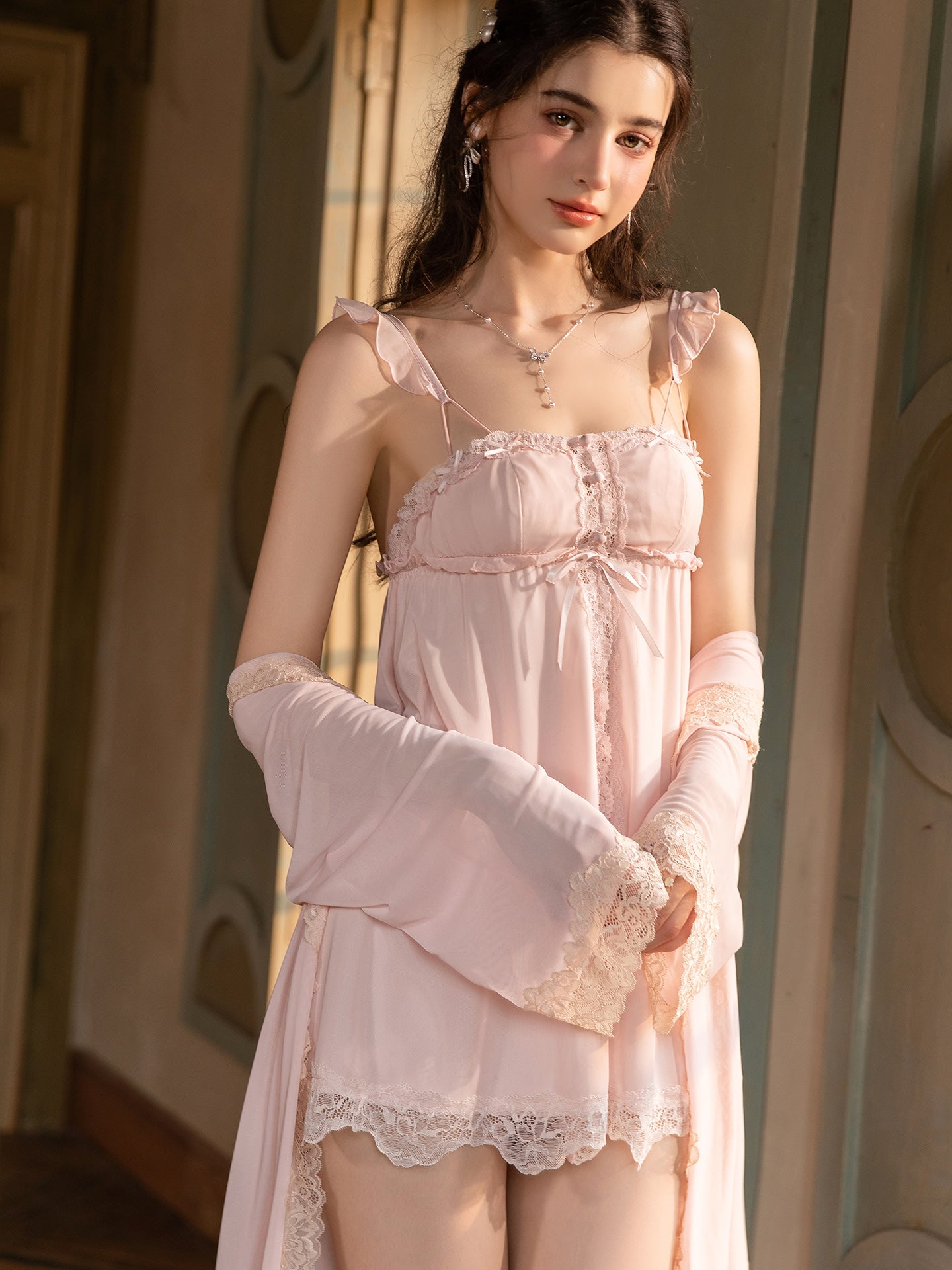 A woman models a light pink lingerie set consisting of a ruffled camisole nightgown and matching long gloves, set against a warm, vintage-style interior background.