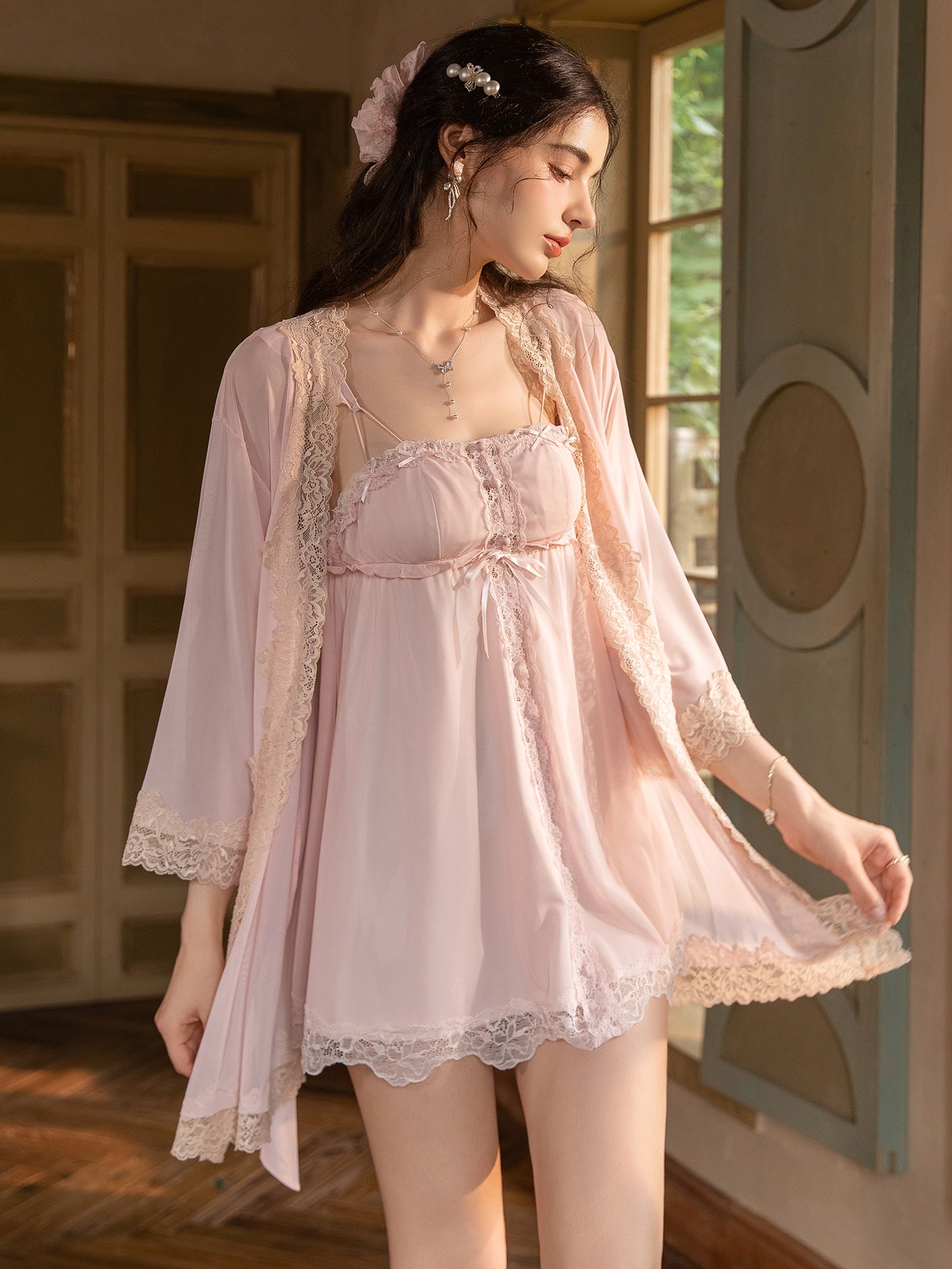 A woman wearing a light pink lingerie set with a matching sheer robe, standing in a softly lit room with warm wood and pastel walls.
