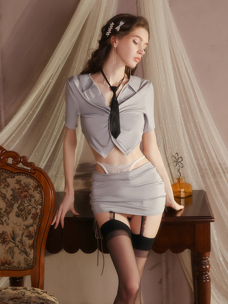 Woman in a gray outfit with a tie standing in a room with curtains and a table.