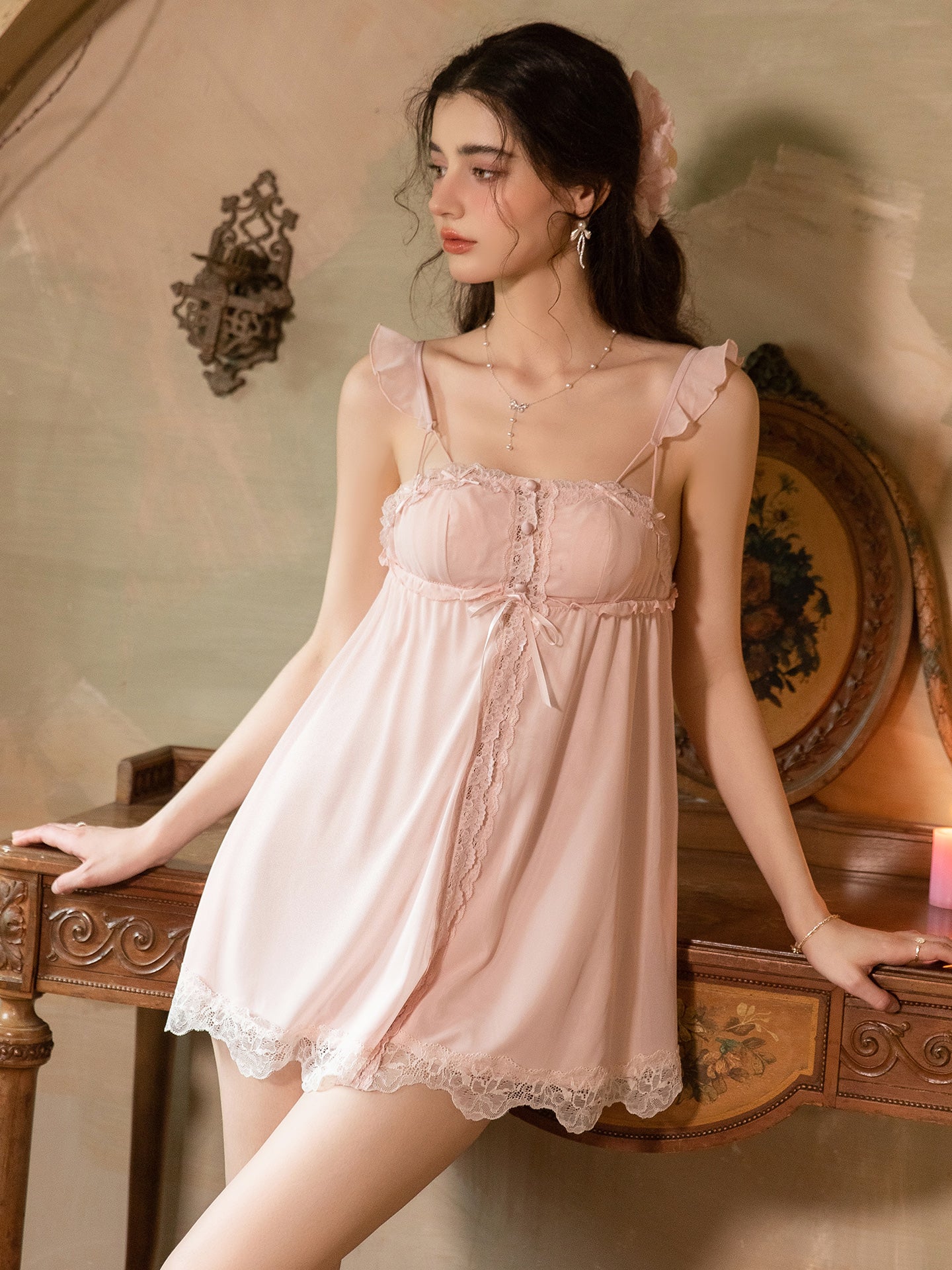 A woman wearing a light pink satin slip dress with lace trim and ruffled shoulder straps, leaning against an ornate wooden table in a vintage-style room.