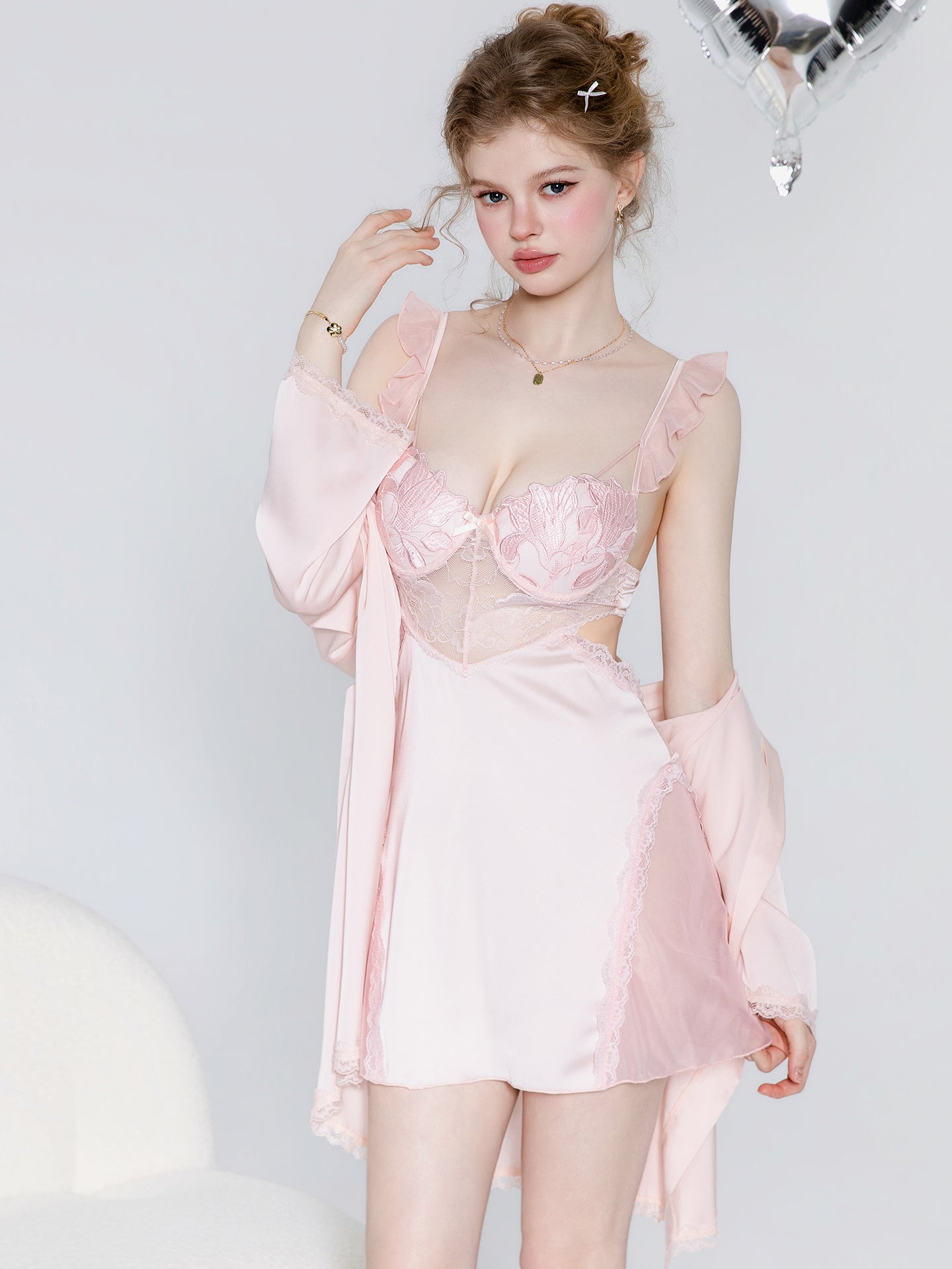 A model wearing a light pink lingerie set consisting of a lace and mesh bustier top, matching satin shorts, and a sheer long-sleeve robe, posed against a light gray background.