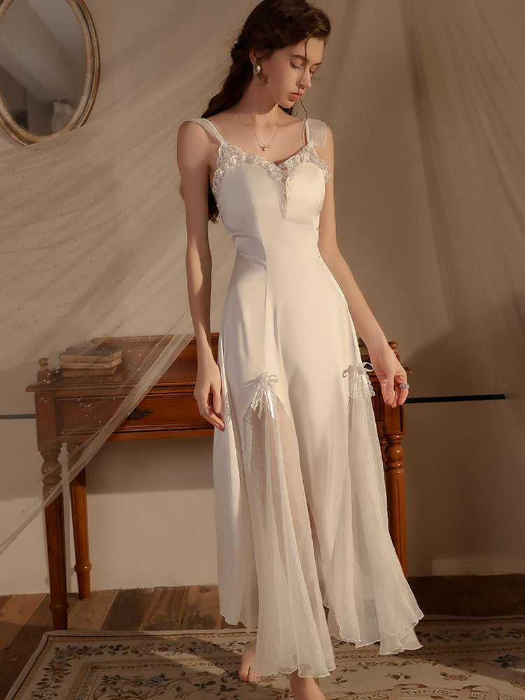 Model wearing a white lace trimmed satin midi slip dress with adjustable straps, semi sheer panels, and bow details, standing by a wooden desk.