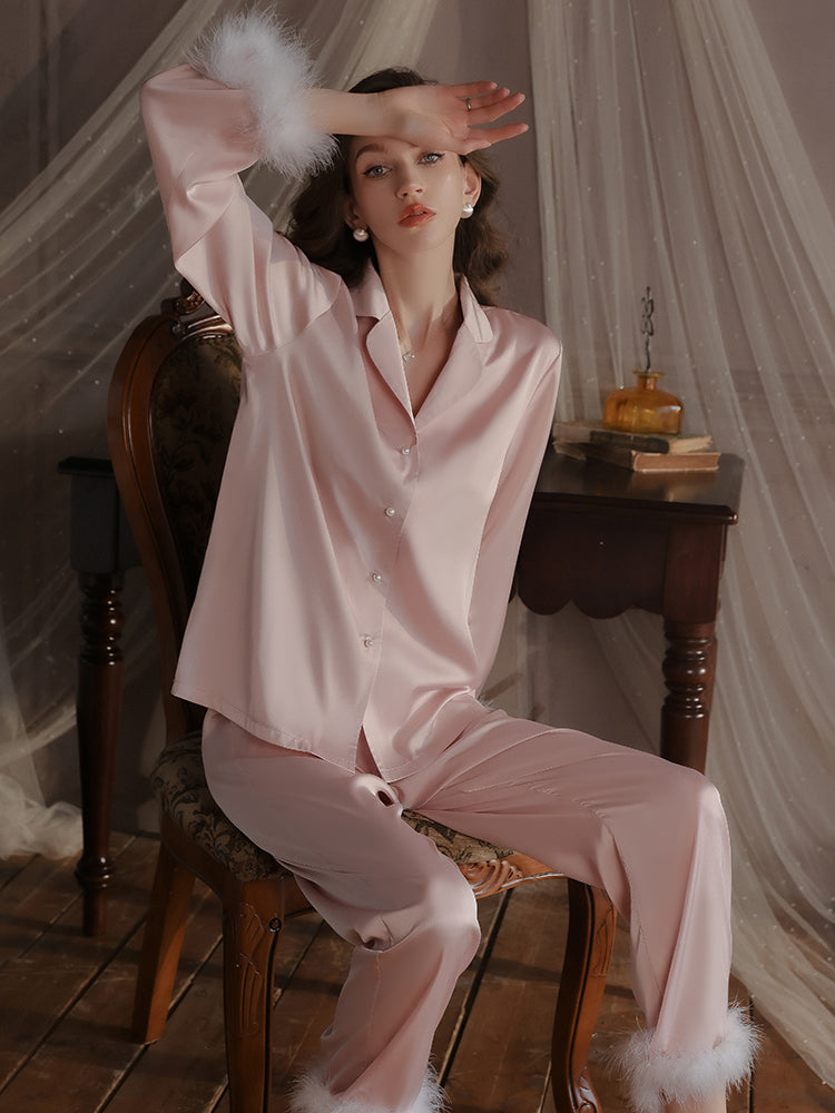 Pink Feather Cuff Satin Pajama Set seated on a chair, featuring the button front top, long sleeves, and matching pants with a relaxed fit.