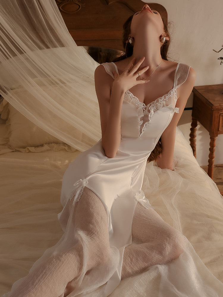 Model sitting gracefully on a bed, showcasing the white lace-trimmed satin slip dress with semi-sheer panels and an elegant flowy hemline.