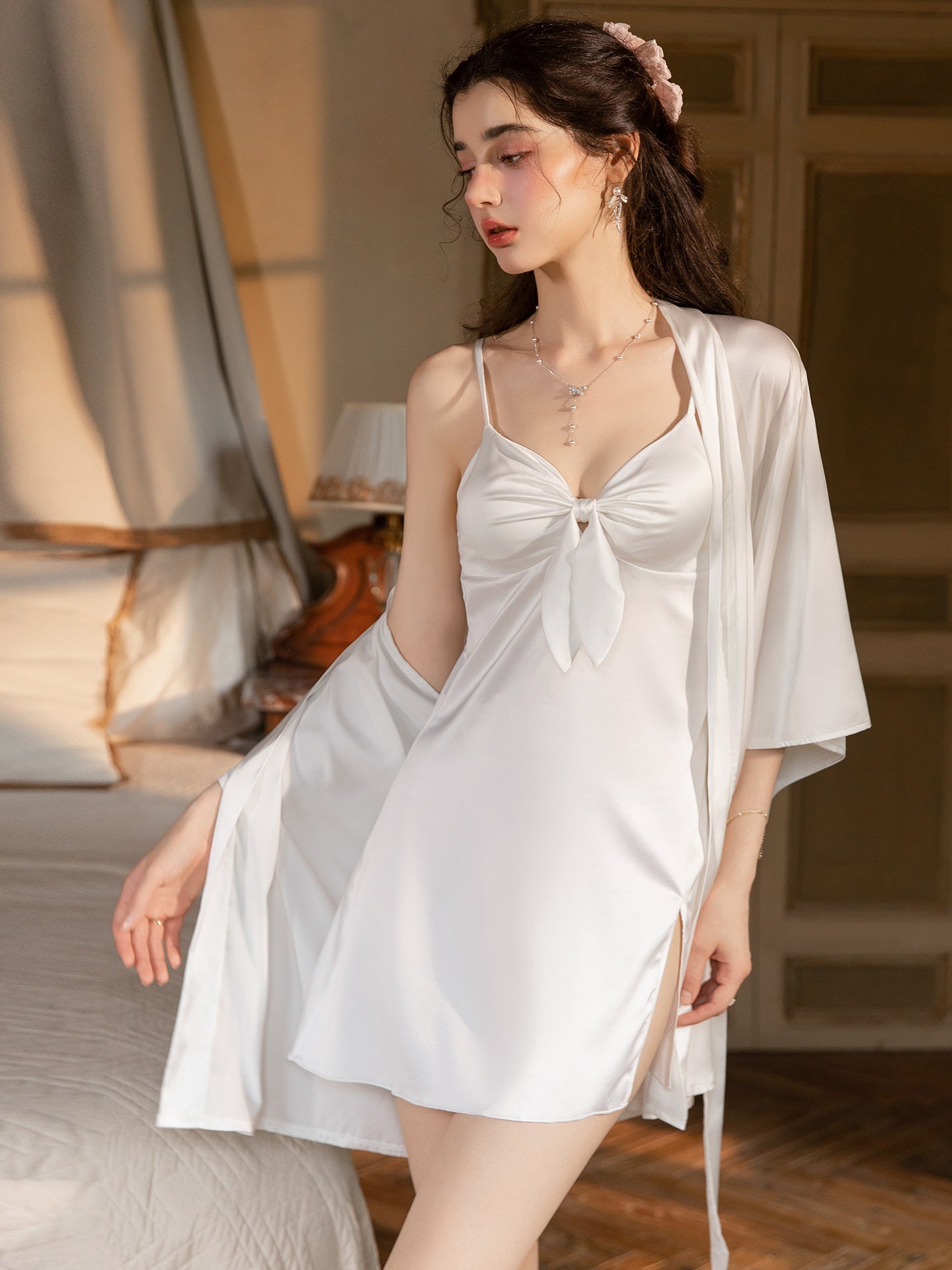 A woman models a white satin slip dress with a front bow detail, paired with a matching white sheer robe, in a softly lit bedroom setting.