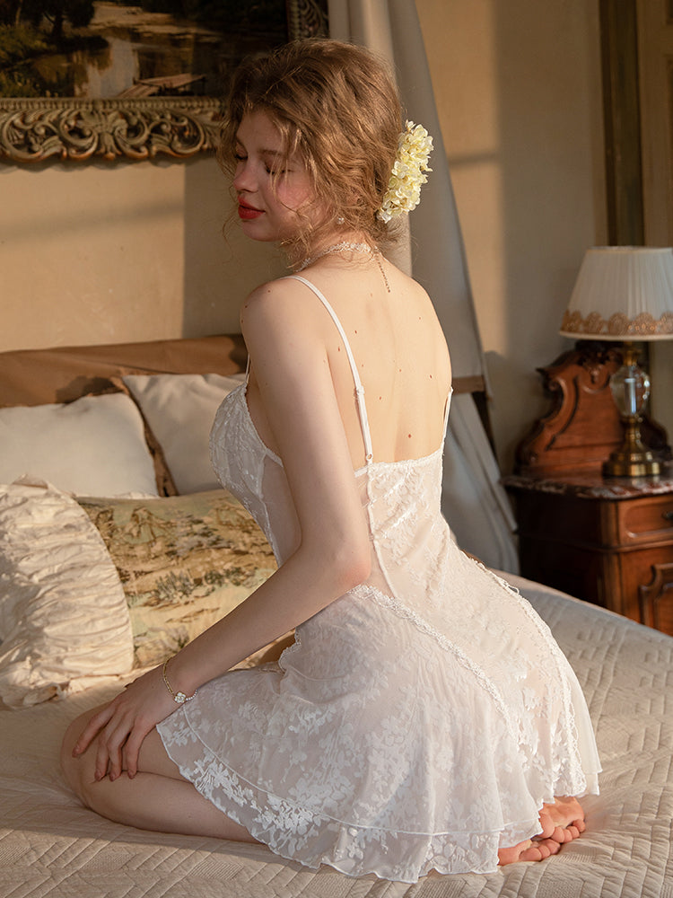 Woman in a lace nightgown sitting on a bed in a softly lit room.white babydoll dress, mini dress, floral embroidered dress, sheer lingerie, white lingerie, chemise nightgown, mini slip dress