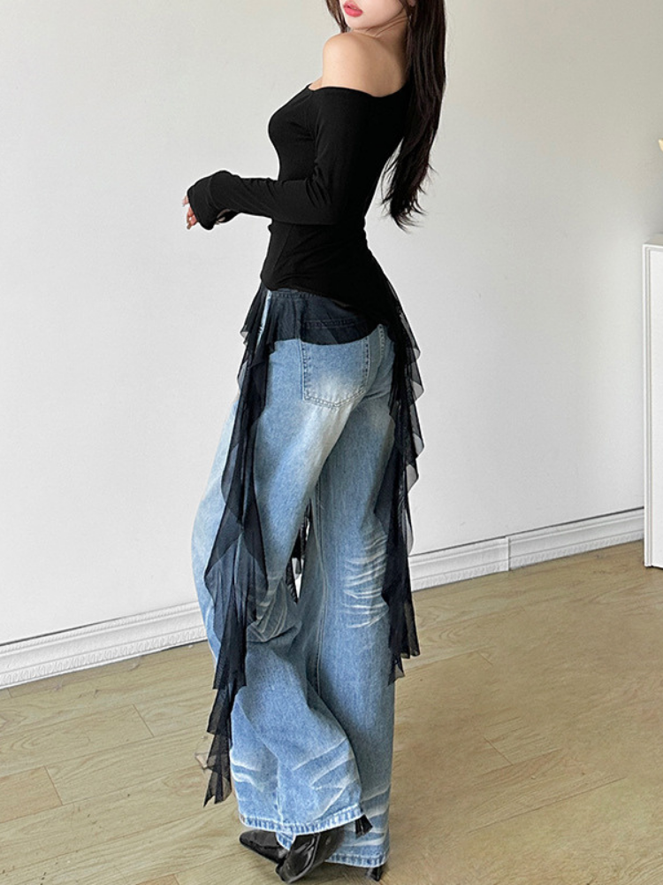 Person wearing a black top and blue jeans with fringe details in a minimal indoor setting.