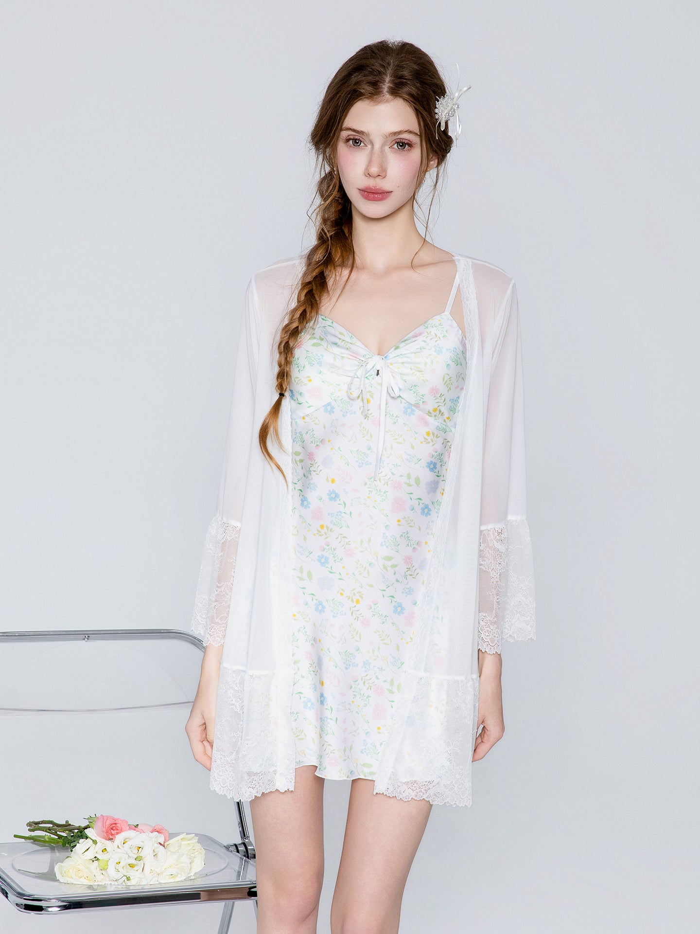 A woman models a white floral nightgown with a sheer white lace-trimmed robe draped over her shoulders, set against a light gray studio background.