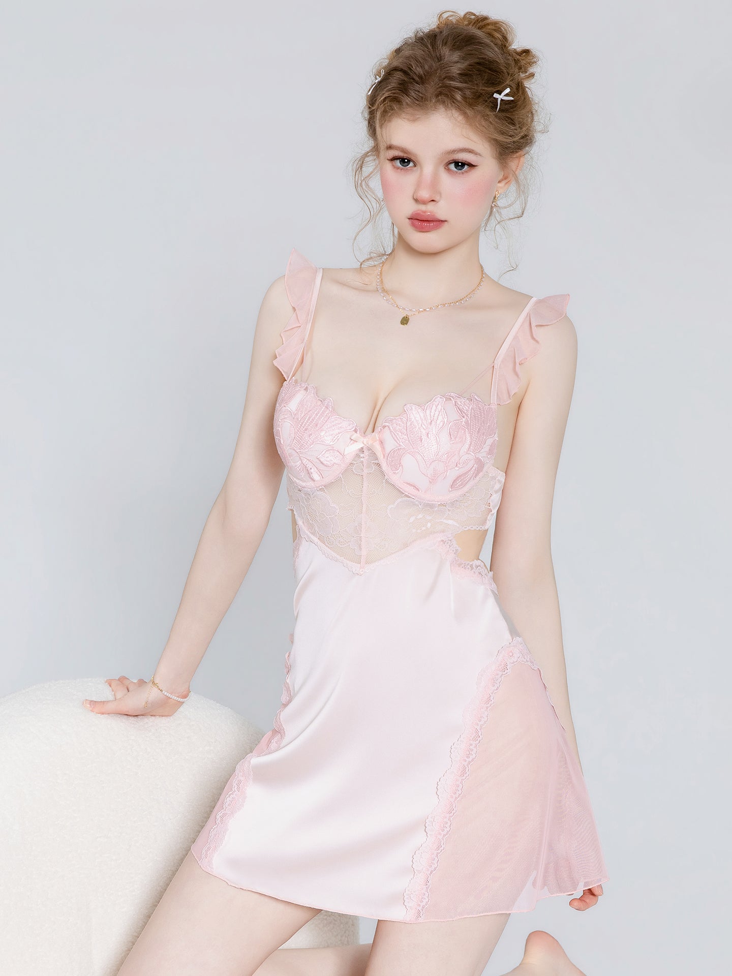 A young woman models a light pink lingerie chemise featuring a lace corset-style bodice with ruffle straps, sheer mesh panels, and a satin skirt, posed against a light gray studio background.