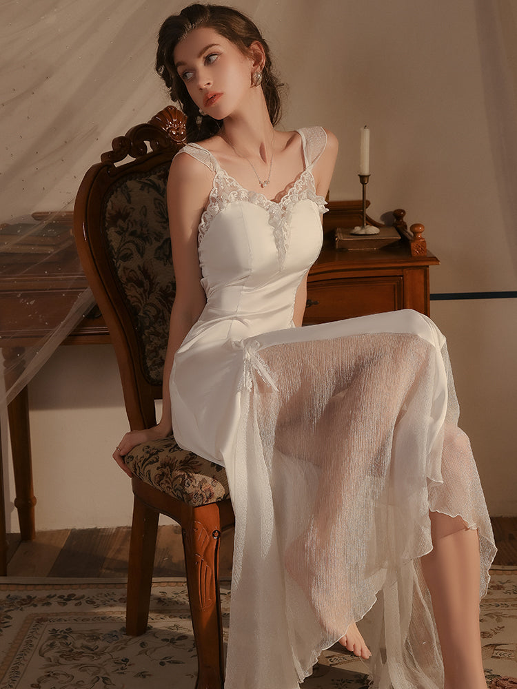 Model sitting on a vintage chair, wearing a white lace trimmed satin midi slip dress with sheer side panels and delicate bow details, accentuating the flowing silhouette.