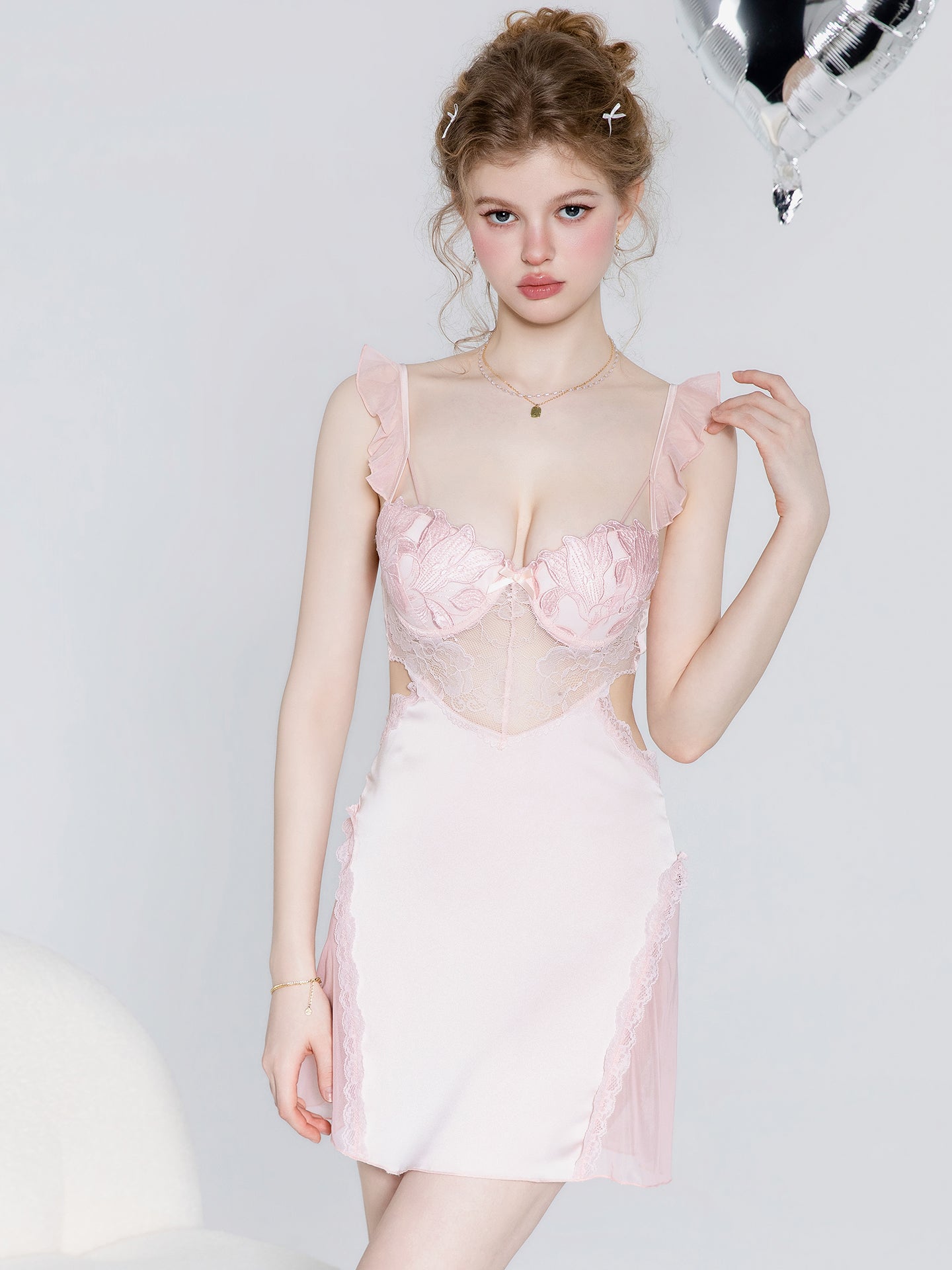 A young woman models a light pink mini dress with a sheer lace corset-style bodice, ruffled shoulder straps, and matching lace side panels, posed against a light gray studio background.