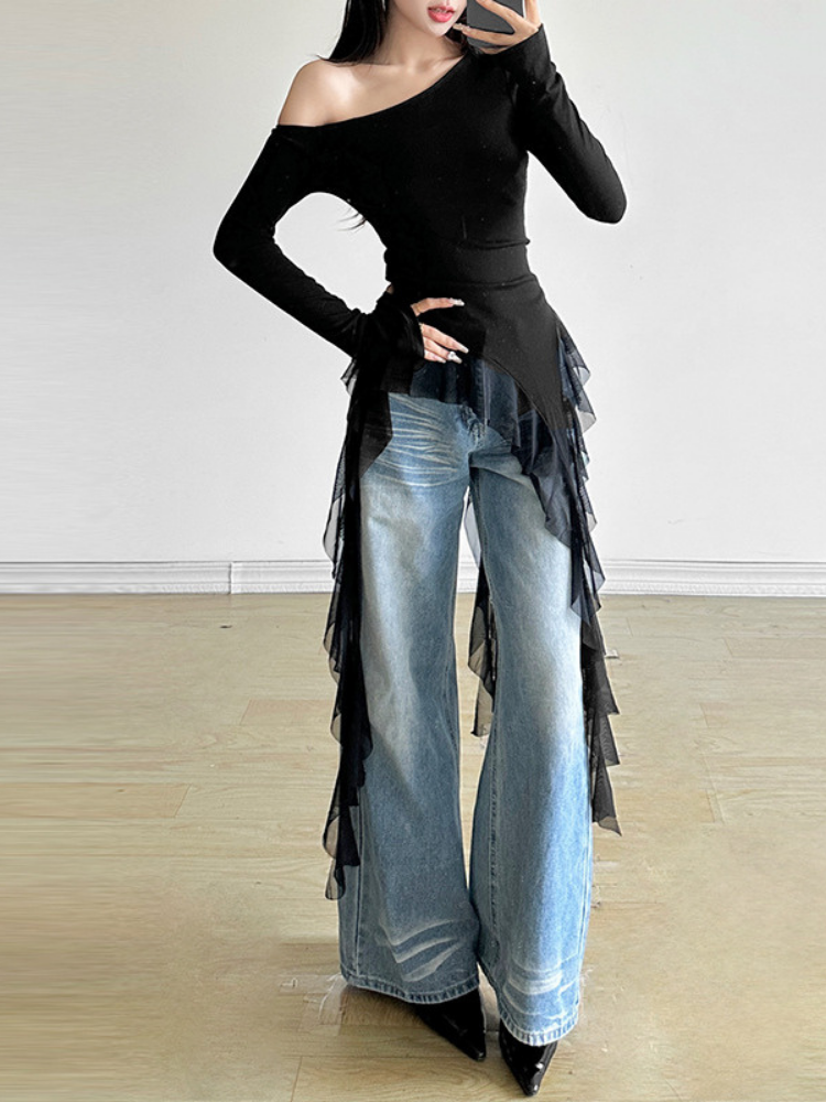 Person wearing a black off-shoulder top with fringe details and light blue jeans indoors.