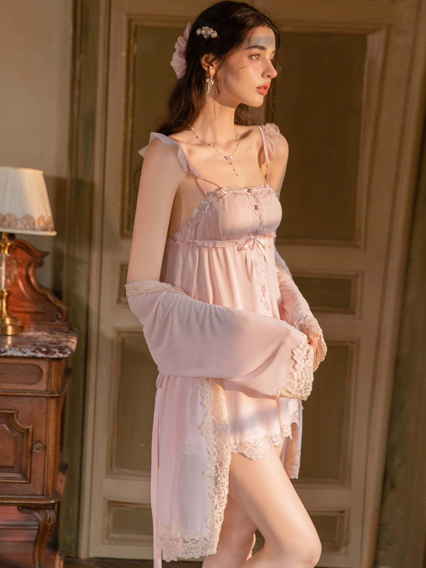 A woman wearing a light pink lingerie set with a matching robe, standing in a warmly lit room with a wooden door and a lamp in the background.