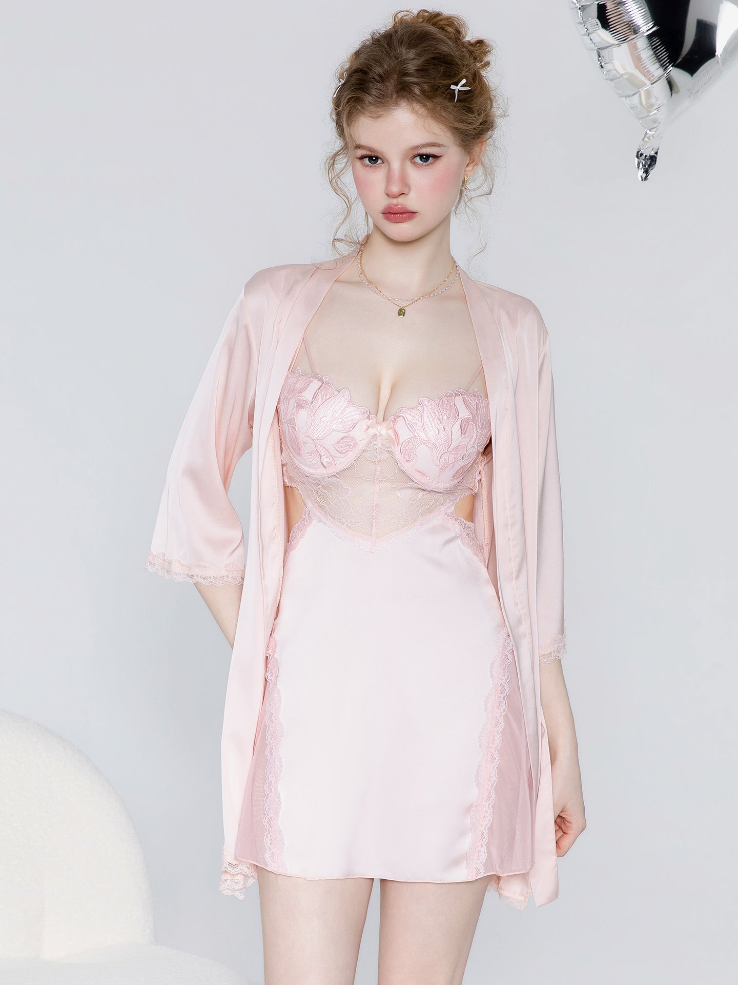 A woman modeling a light pink lingerie set consisting of a satin robe and a matching lace-trimmed slip dress against a plain light gray background.